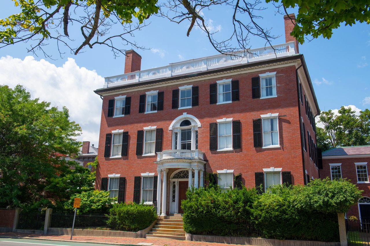 Red-brick building with black shutters, a columned entrance, and rooftop balustrade behind leafy hedges.
