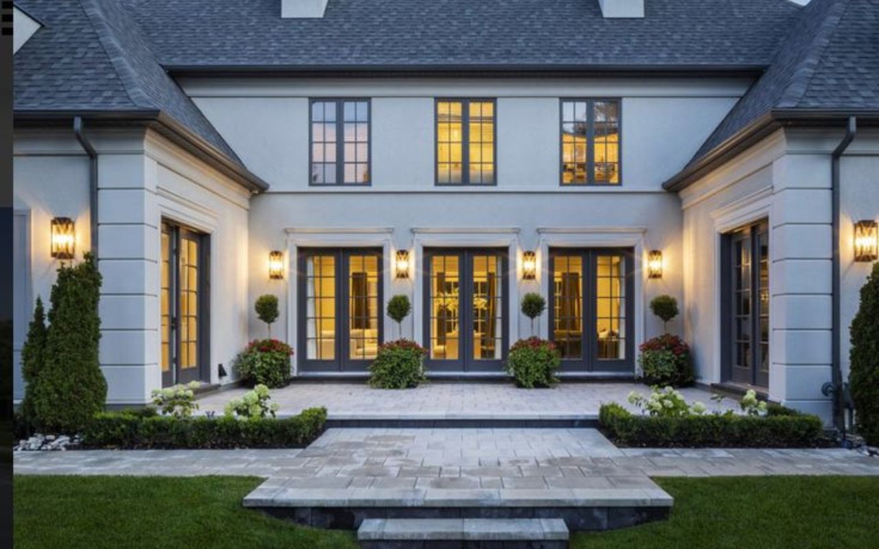 White house with warmly lit windows, a stone terrace, and formal landscaping at dusk.