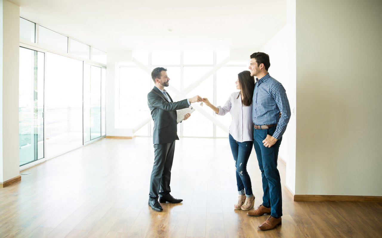 Real estate agent hands keys to a couple in a bright, empty home with large windows.