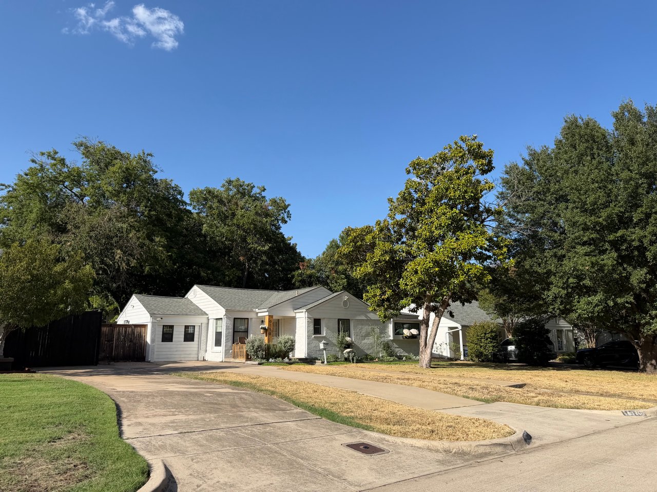 White single-story house with a broad concrete driveway and mature trees along a residential street.