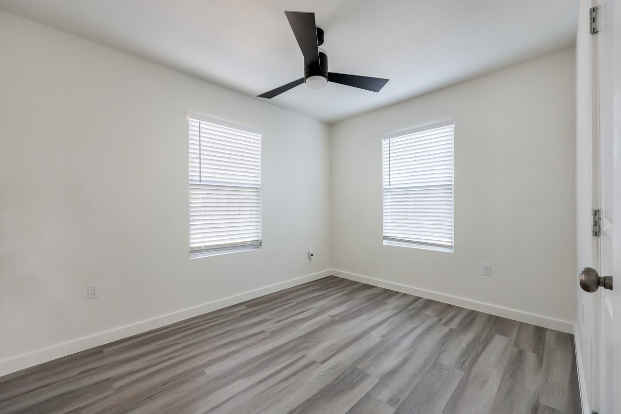 Empty bedroom with gray plank flooring, two windows with blinds, and a black ceiling fan.