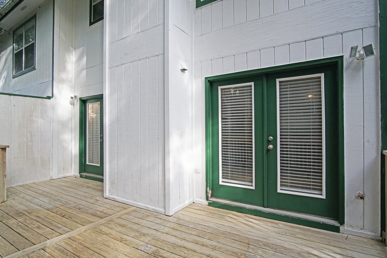 Back deck at 529 Collinsford Road, Tallahassee, Florida, featuring two sets of French doors with green trim and glass panels with blinds. The area has light-colored wood decking and white vertical siding, creating a clean, open space for outdoor relaxation or entertaining.