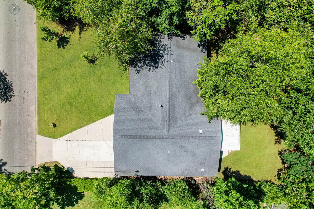 Aerial view of a house with a dark gray roof surrounded by green trees. There's a driveway leading to the road on the left, adjacent to a small grassy yard.