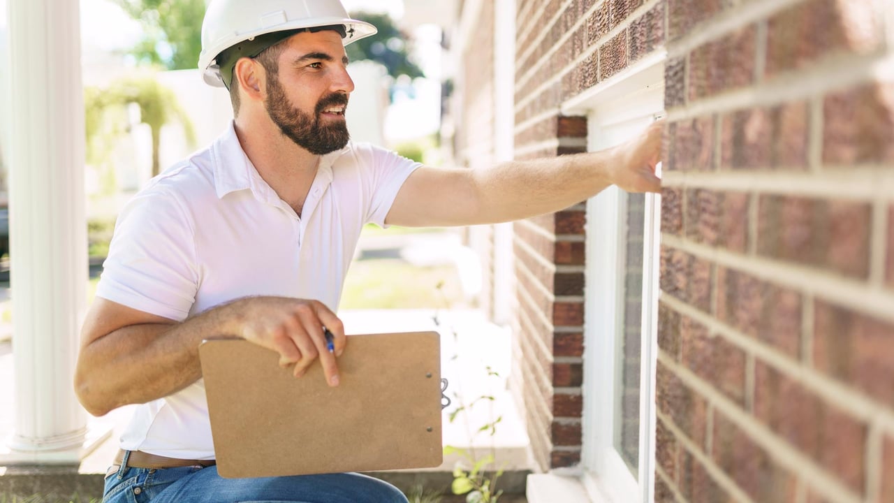 A licensed home inspector wearing a hard hat and holding a clipboard inspecting an exterior basement window and brick foundation, illustrating the smart risk management strategies detailed in our home inspection guide for GTA buyers and sellers