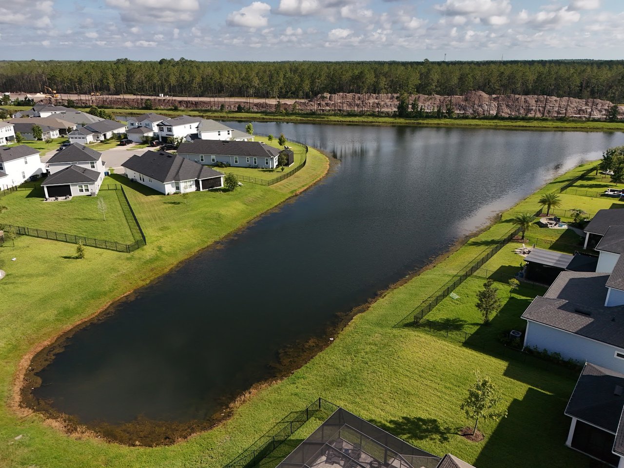 Aerial view of homes with fenced yards along a lake, with a wooded horizon beyond.