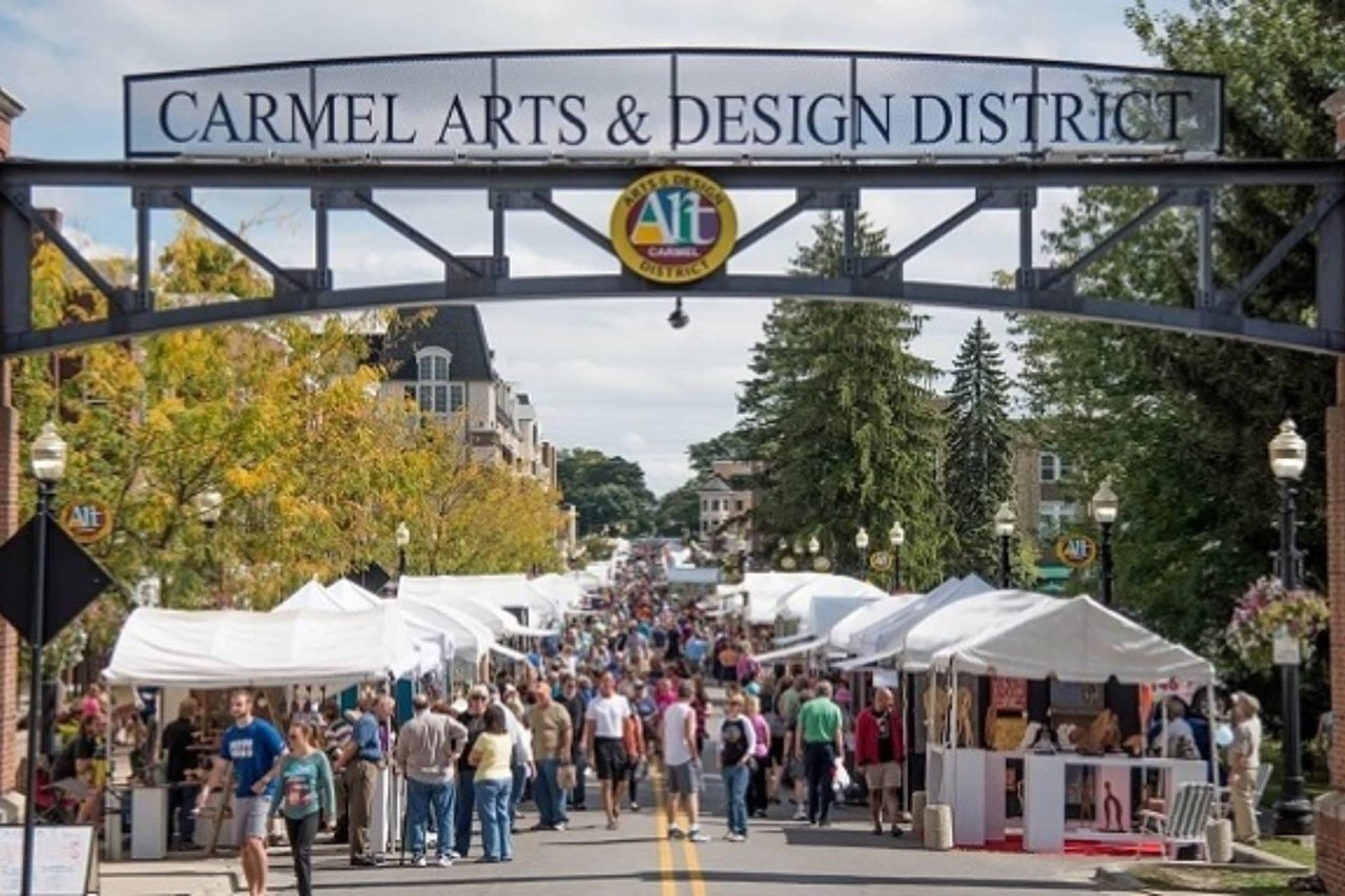 An outdoor community art fair in the Carmel Arts & Design District featuring rows of artisan booths, white vendor tents, and festival crowds.