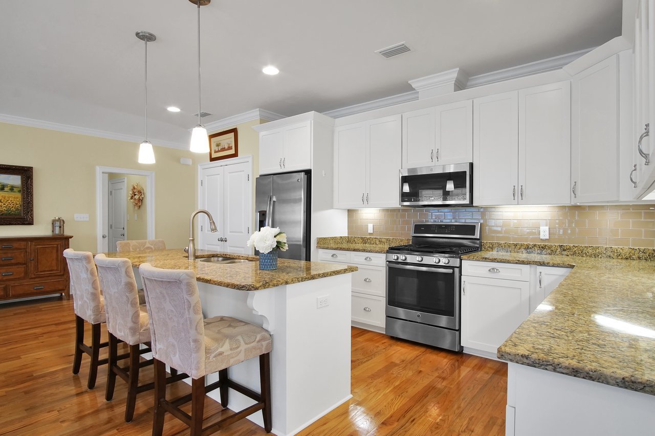 Bright kitchen with white cabinets, granite countertops, and an island with three upholstered stools. Stainless steel appliances and hardwood floors.