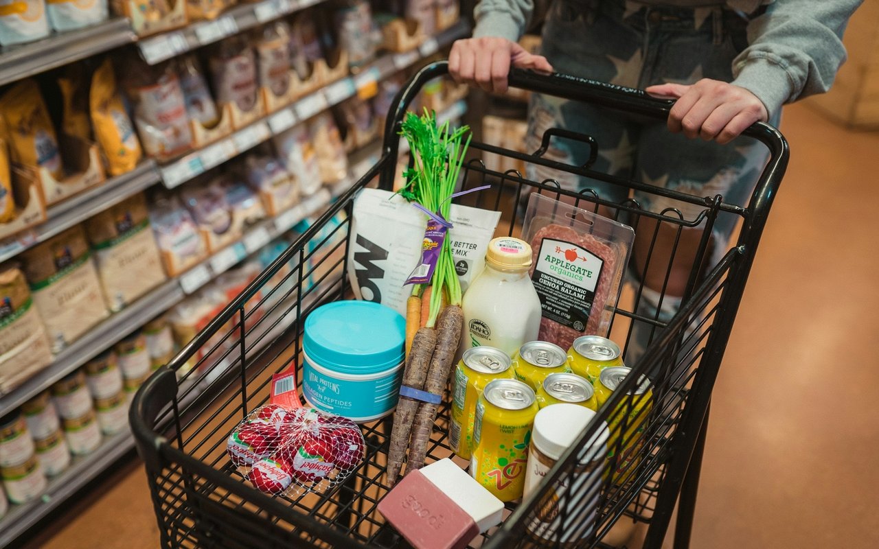 A grocery cart filled with produce, canned drinks, and packaged foods in a supermarket.