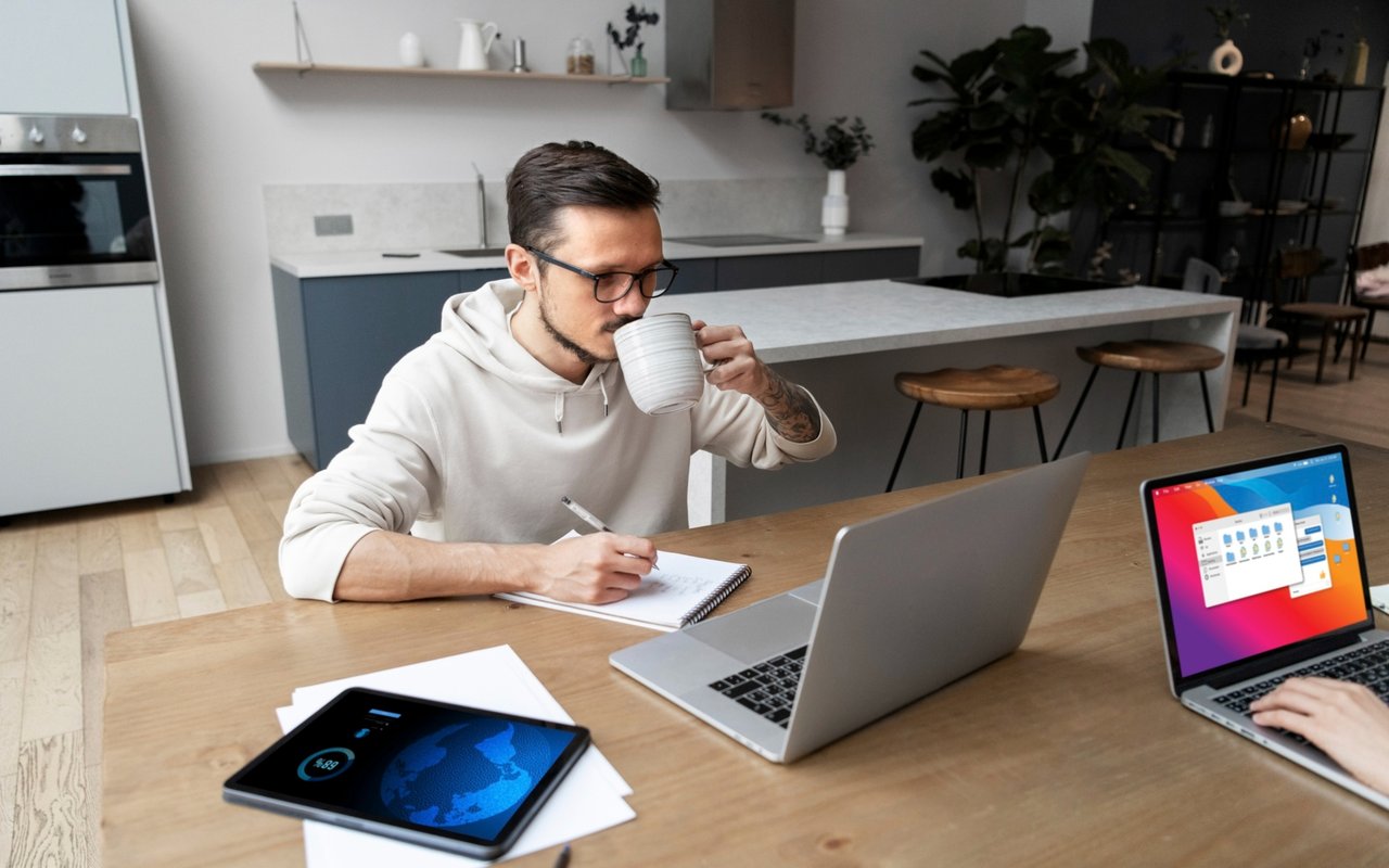 Man working remotely at a kitchen table, writing in a notebook beside a laptop and coffee.