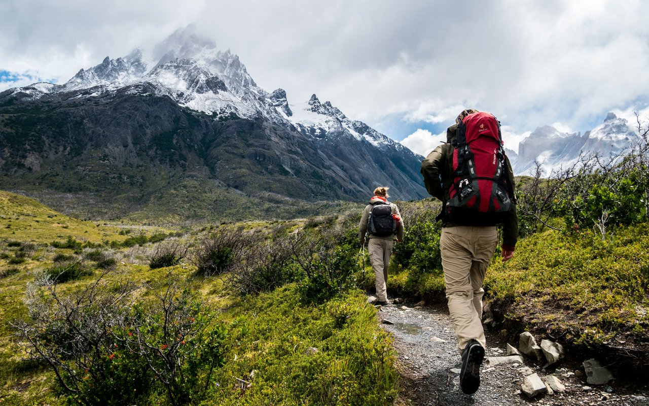 Two hikers follow an alpine trail toward snow-covered mountains.