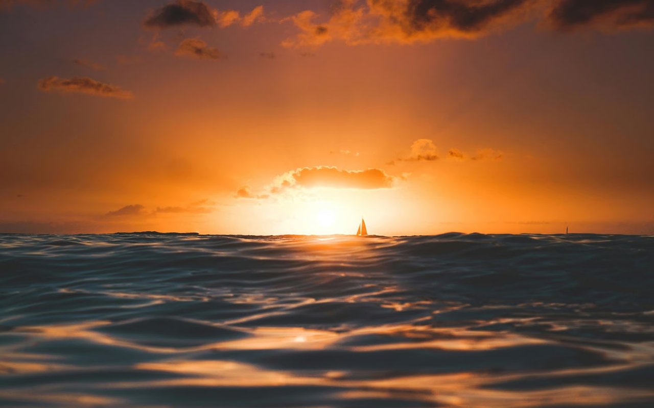 Sailboat on the horizon at sunset, with orange light reflecting across the ocean.