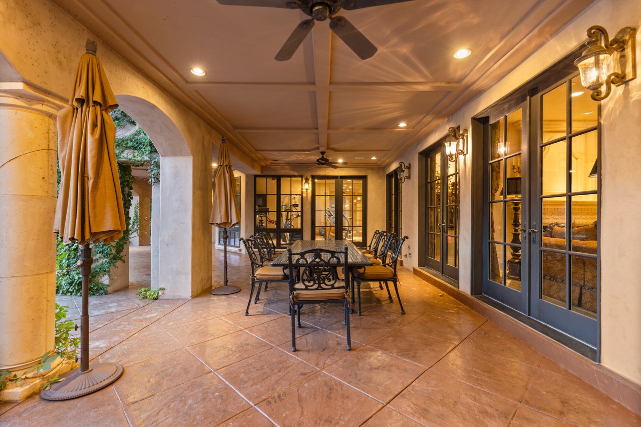 Covered patio with a long dining table, ornate chairs, stone tile, and black-framed glass doors.