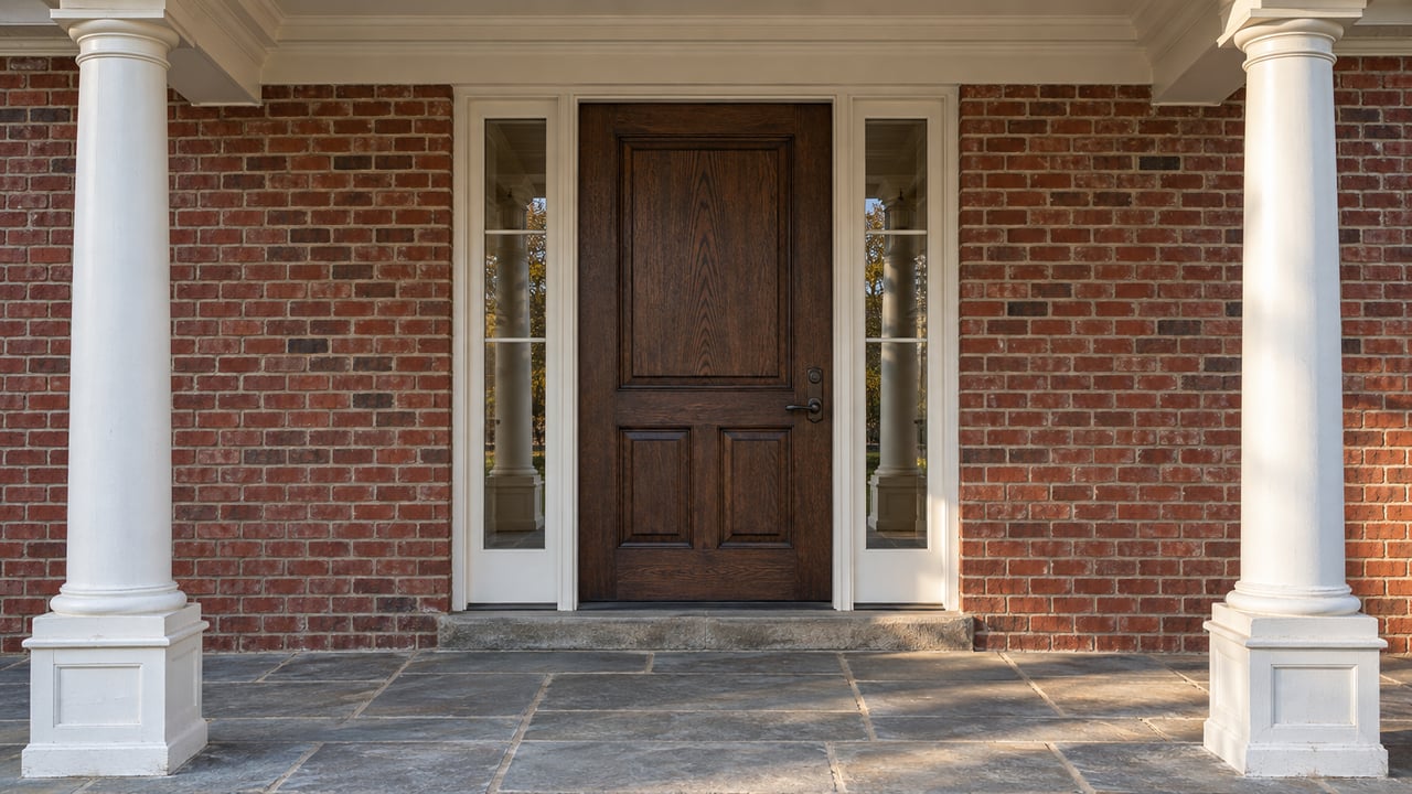 Paired white columns frame a closed dark oak front door and glass sidelights in a red brick entry, with worn gray-blue stone underfoot.