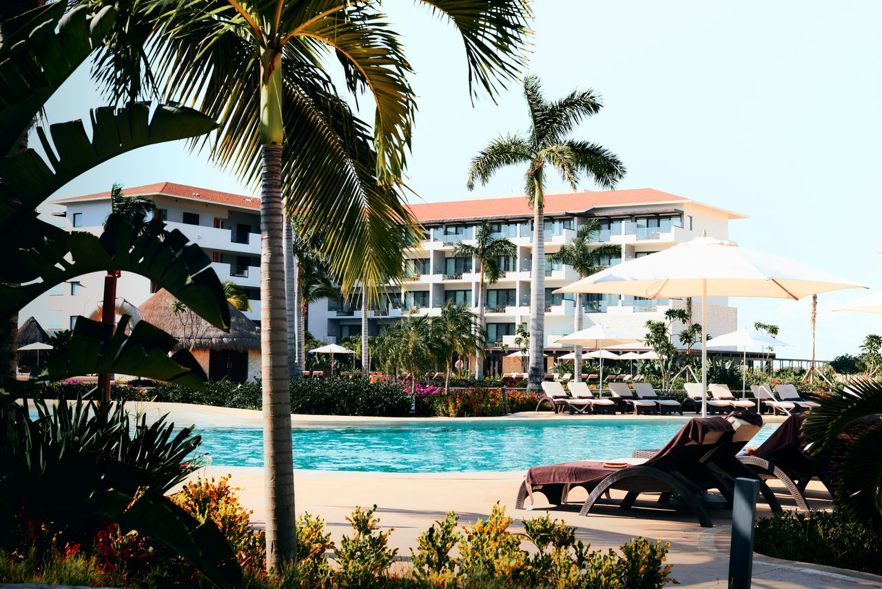 Scenic view of a resort pool surrounded by tropical palms, sun loungers with umbrellas, and a multi-story building in the background.