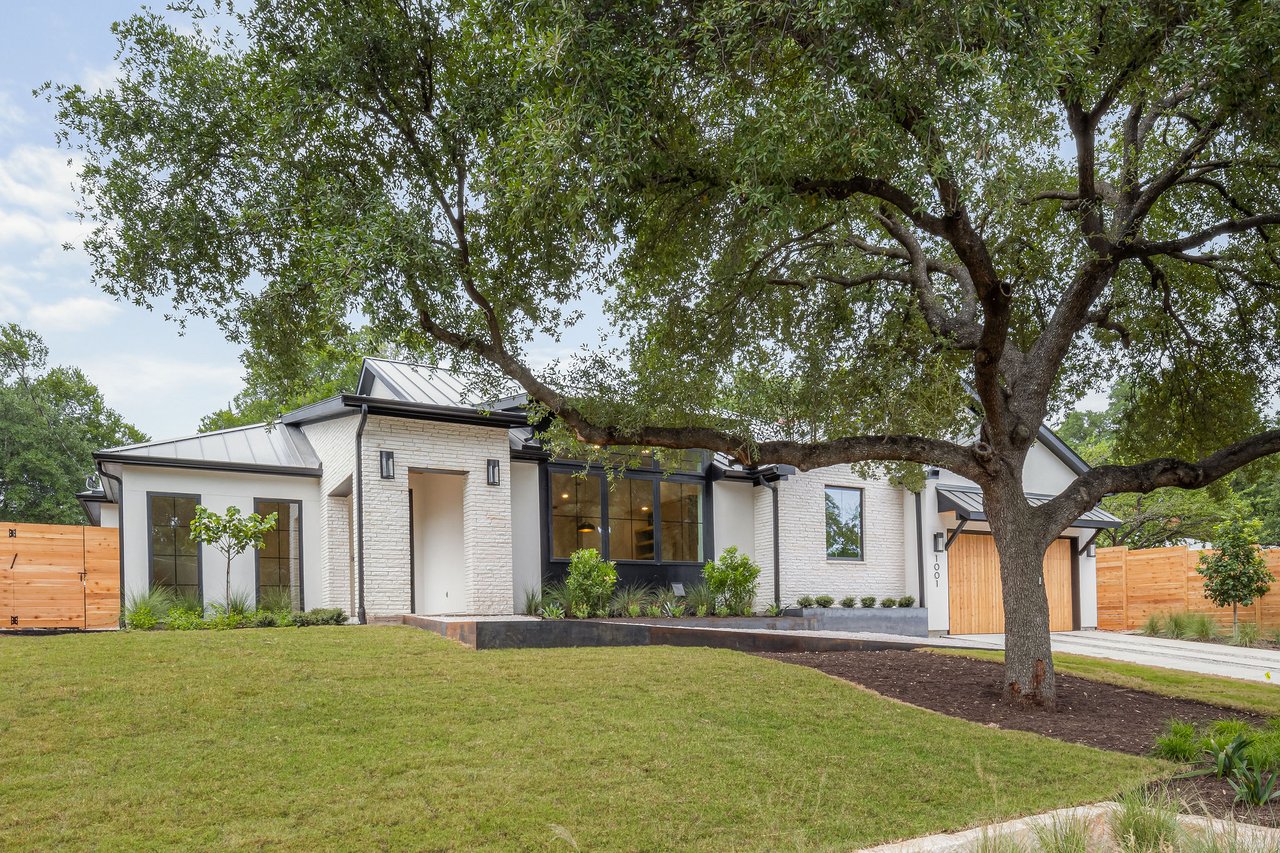 Front elevation view of 1001 Bluebonnet Ln in Zilker, Austin, TX, highlighting the white brick exterior, black-framed windows, and mature oak tree in the front yard. Built by Rivendale Homes.