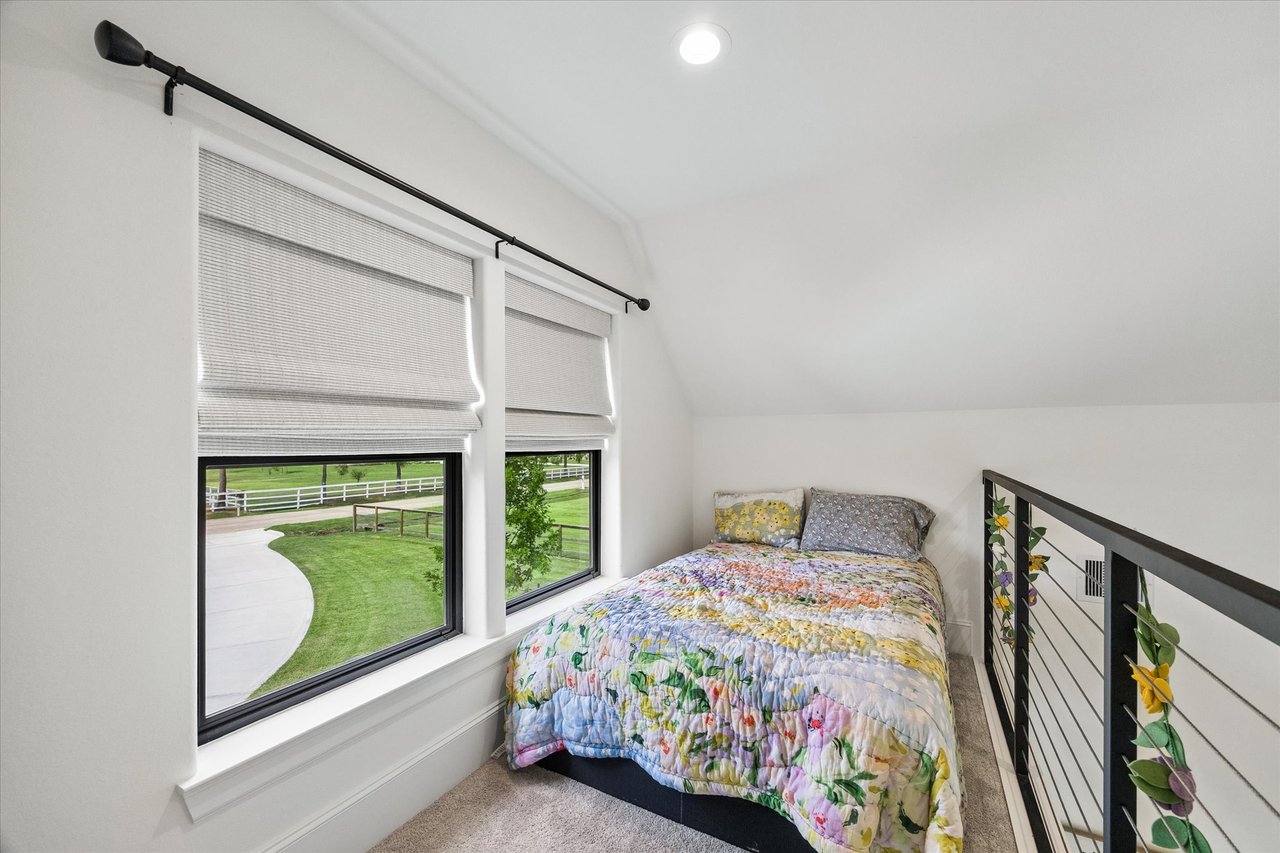 Loft bedroom with colorful floral bedding, large shaded windows, and a black metal railing.