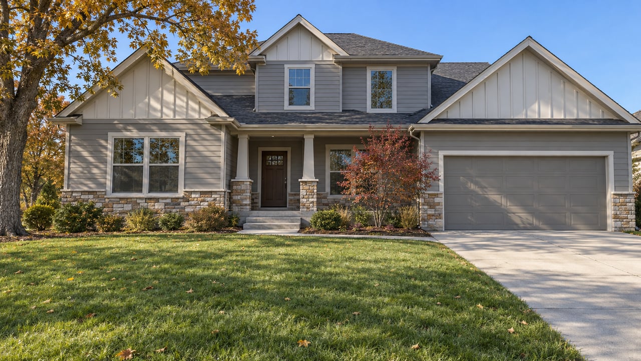 Two-story gray-sided home with stone porch piers, a deep autumn lawn, and a spreading oak under clear blue sky.