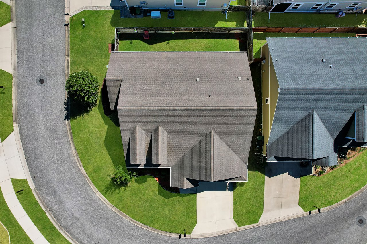 Aerial view of a suburban neighborhood with houses featuring grey roofs, green lawns, and a curved road, conveying a tidy and orderly atmosphere.