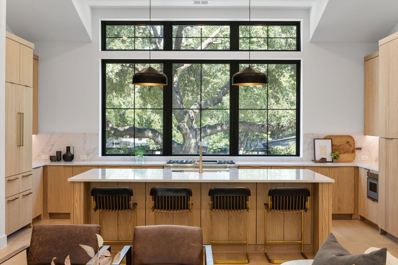 Kitchen at 1001 Bluebonnet Ln in Zilker, Austin, featuring a quartz waterfall island, black pendant lighting, and wood cabinetry, with a wall of windows overlooking the trees.