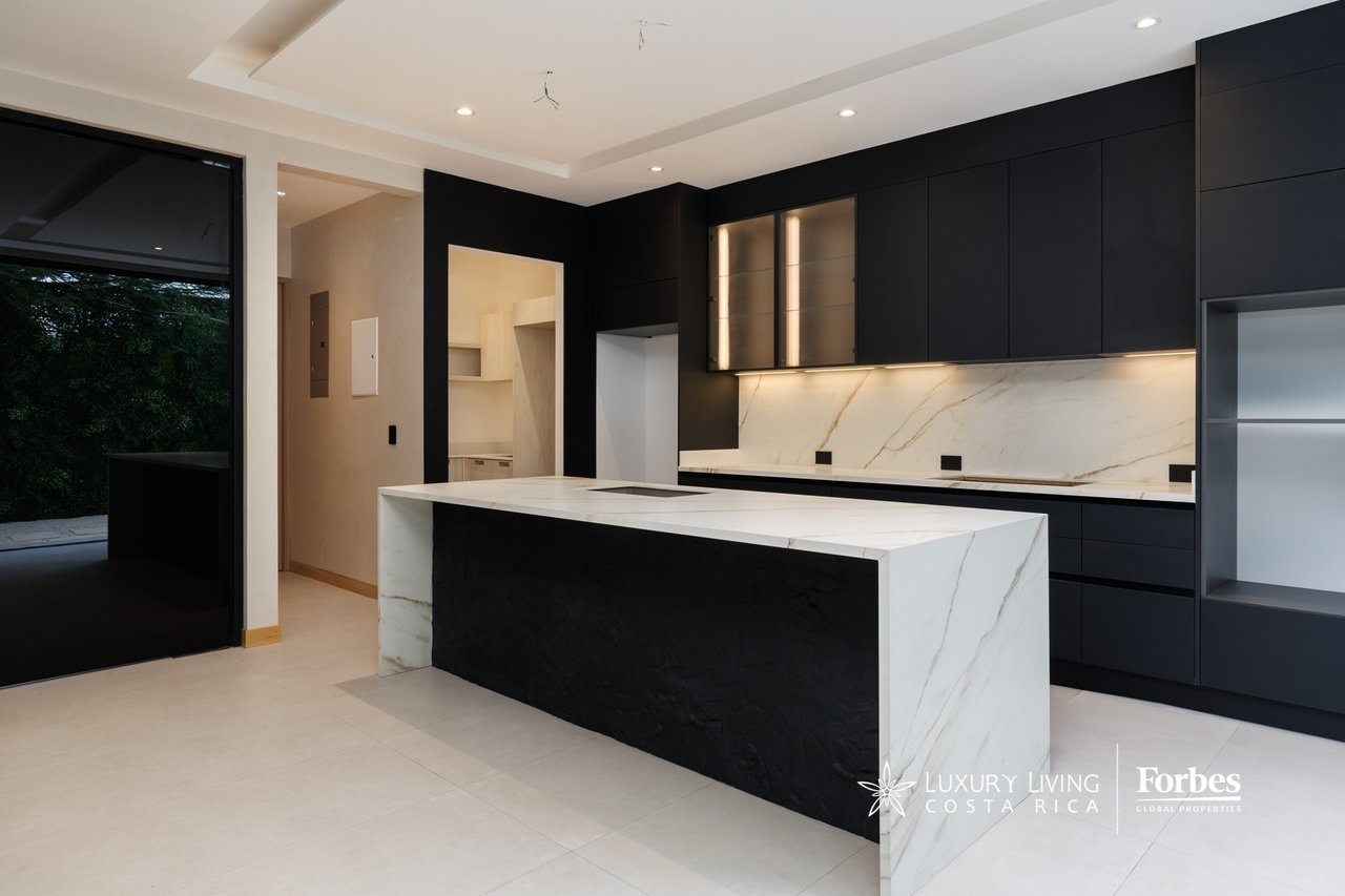 Modern kitchen with black cabinetry, a large marble island, and a glass door opening toward a garden.