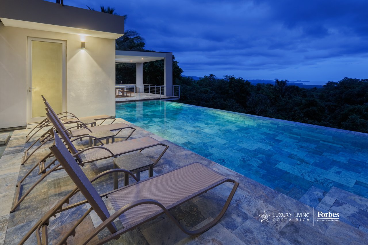 Infinity pool and lounge chairs on a hillside terrace overlooking forest and ocean at dusk.