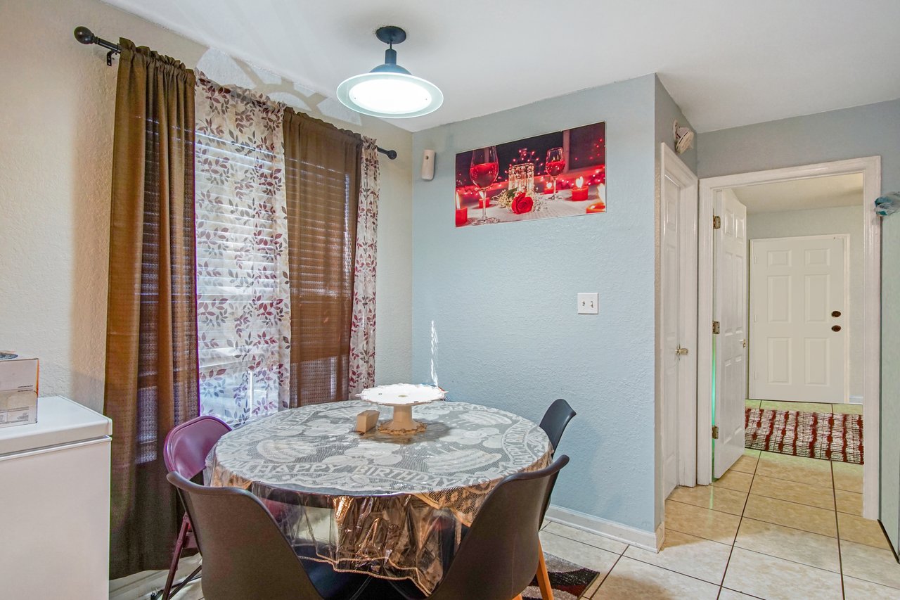 Dining nook at 2506 Nugget Lane in Tallahassee, Florida. The space features light blue walls, tile flooring, and a round dining table with four chairs under a modern pendant light. A wall art piece with wine glasses and candles decorates the wall, while a window with patterned curtains allows natural light into the room.