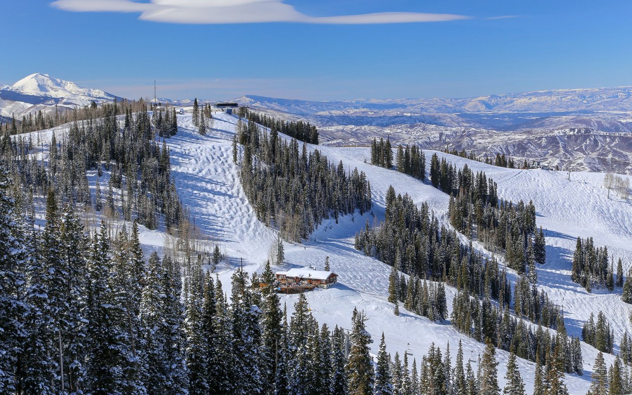 Snowy ski slopes wind through evergreen trees toward a lodge and distant mountain ranges.