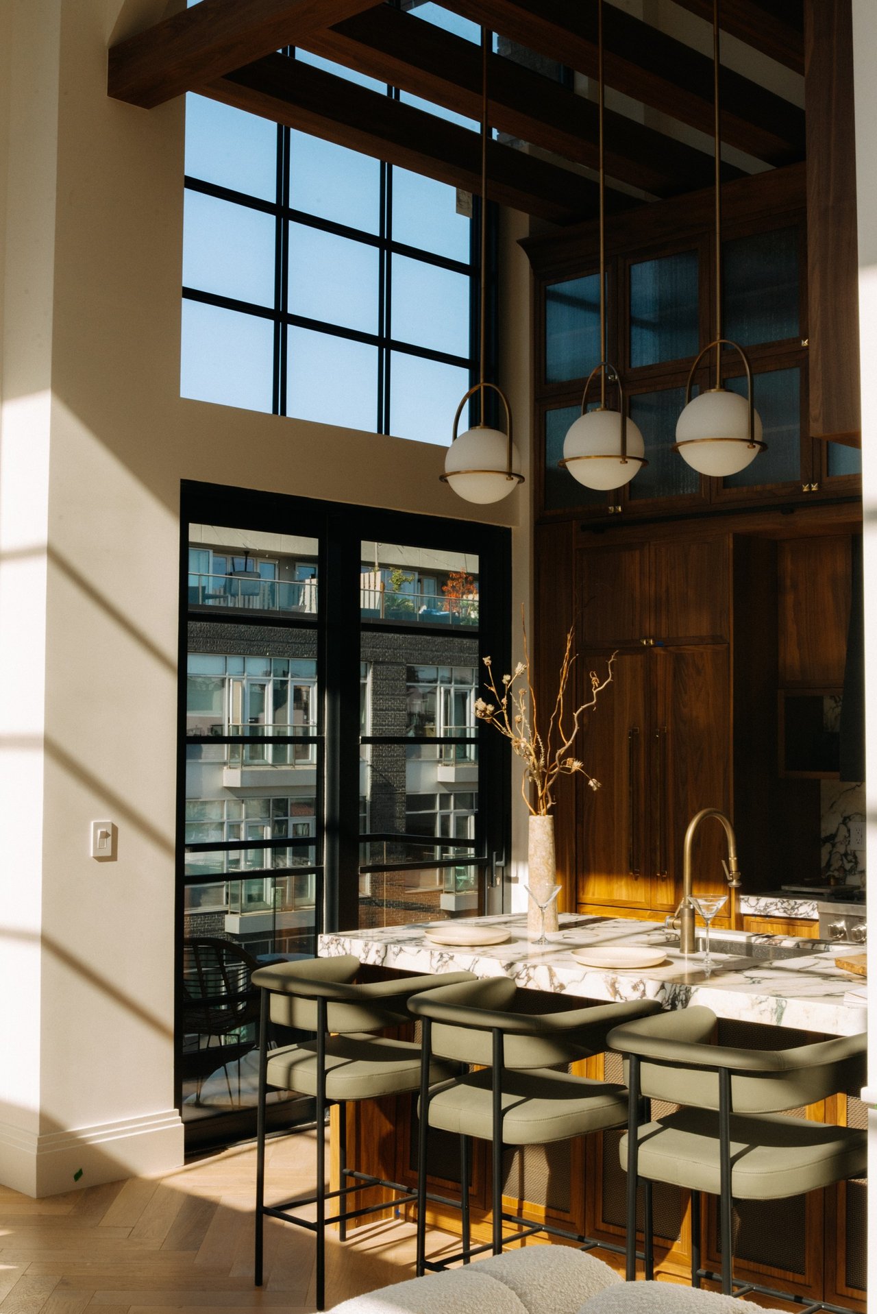 Kitchen with a marble island, dark wood cabinetry, globe pendants, and tall windows beneath exposed beams.