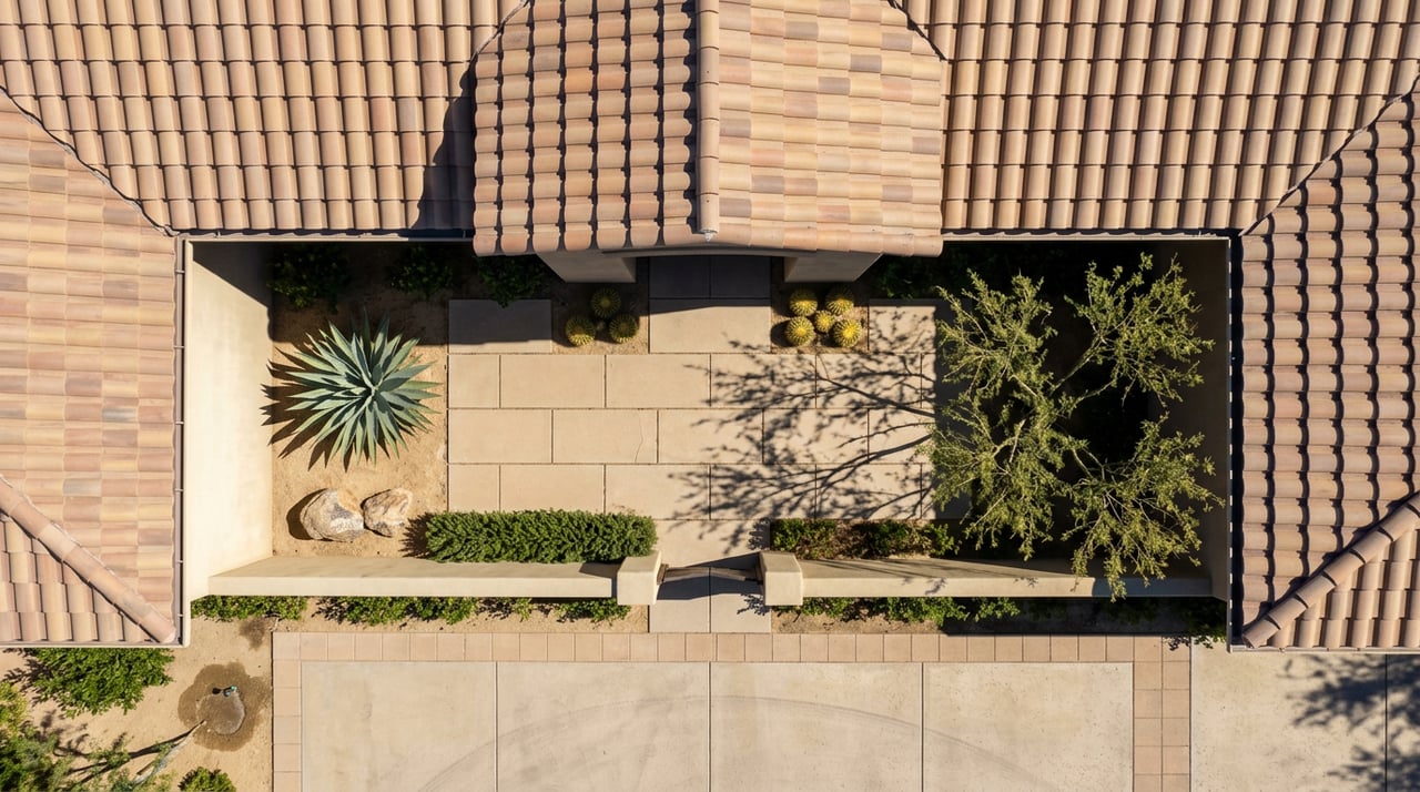 Overhead courtyard with terracotta tile roof, agave, desert trees, and sunlit paving striped by long shadows.