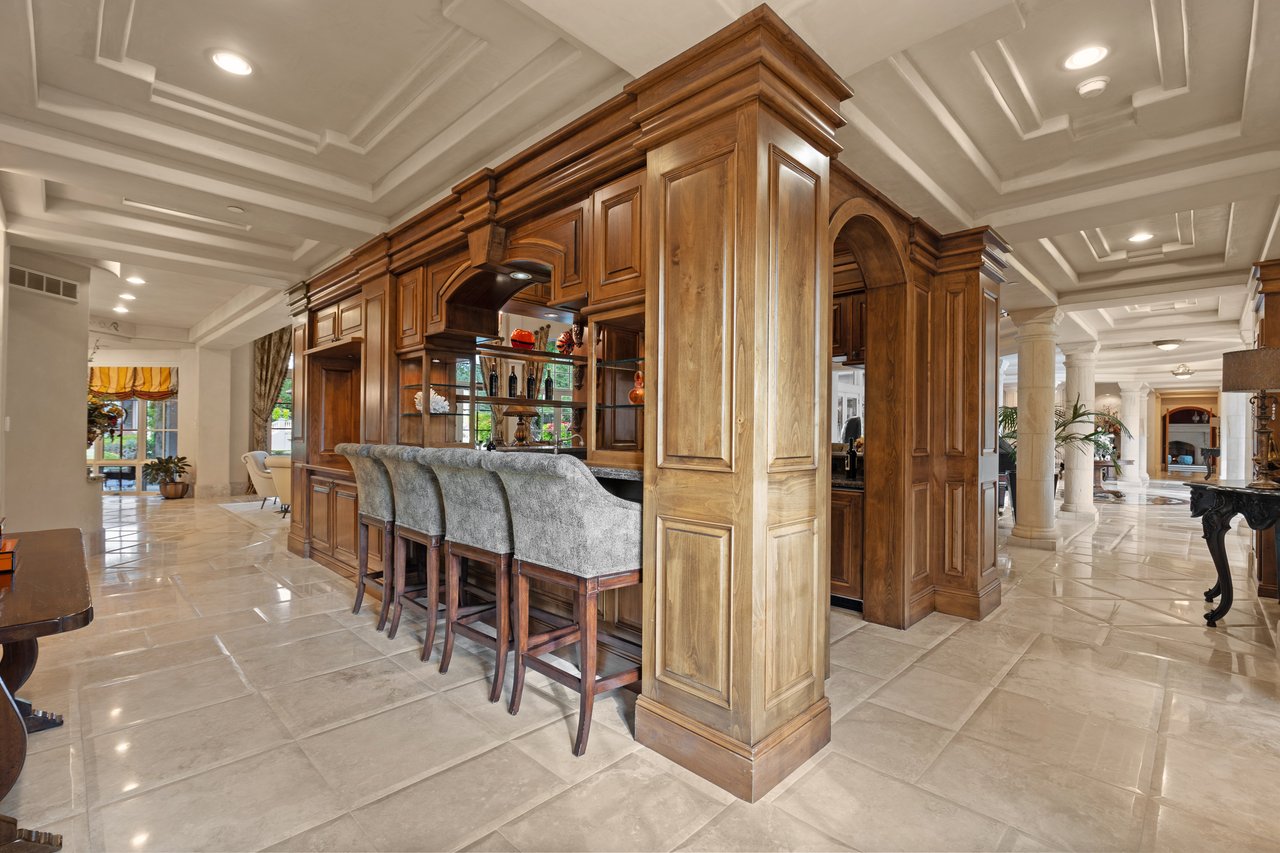 Built-in wood bar with upholstered stools beneath a coffered ceiling in a spacious home interior.