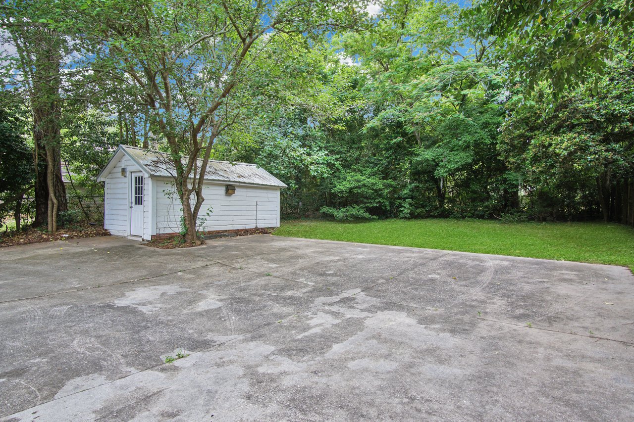 A small white shed sits beside a large concrete area, surrounded by lush green trees and grass. The scene is calm and serene under a clear blue sky.