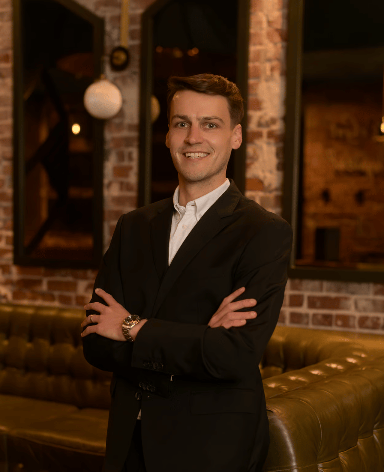 Spencer Griffith in a black suit, arms crossed, in a warmly lit brick-walled lounge.