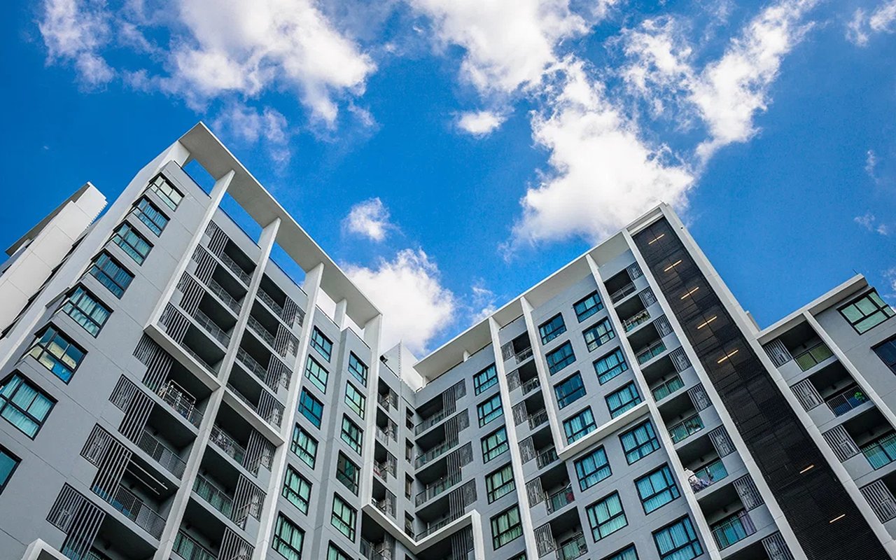 Modern condominium building with white façades and rows of balconies against a blue, cloudy sky.