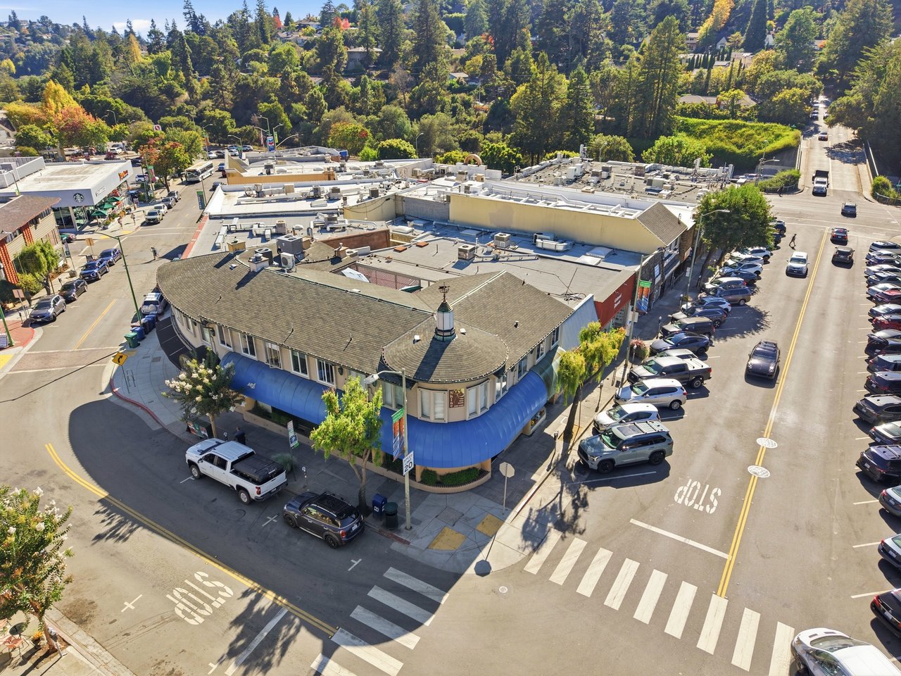 Aerial view of a corner commercial building with blue awnings, surrounding streets, and a wooded hillside.