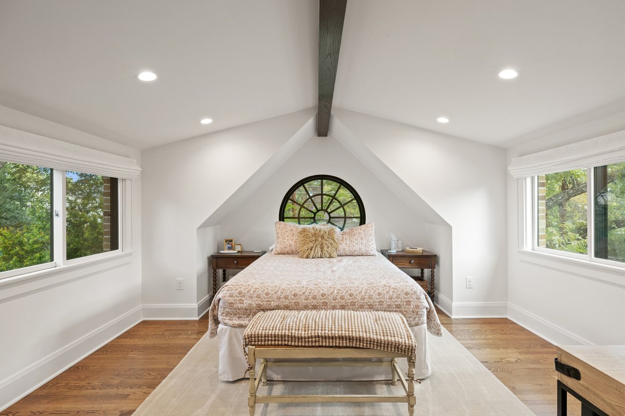 Staged bedroom with a round window above the bed and an exposed ceiling beam.