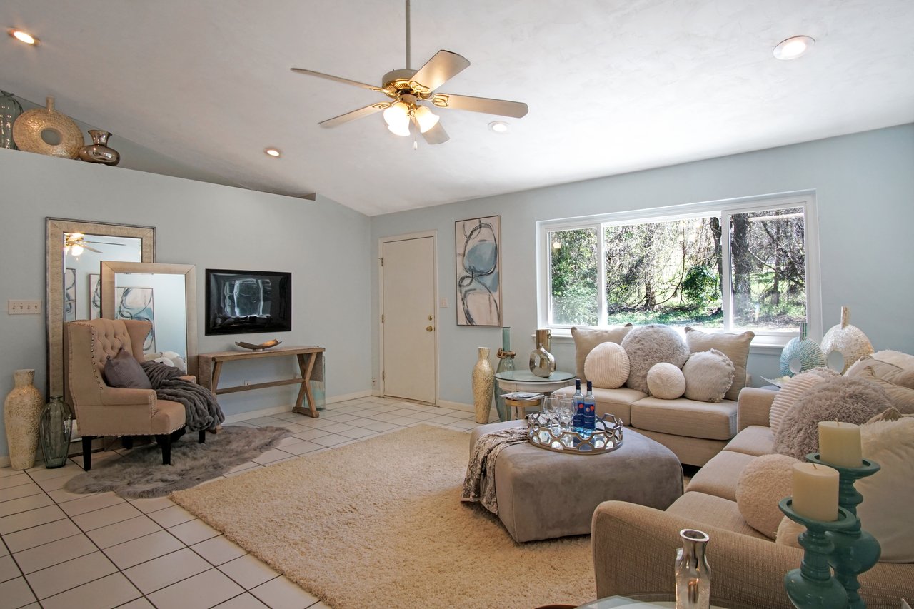 Cozy living room with soft beige and blue tones. Plush sofas adorned with round pillows, a large window revealing lush greenery outside, exudes calmness.Cozy living room with soft beige and blue tones. Plush sofas adorned with round pillows, a large window revealing lush greenery outside, exudes calmness.