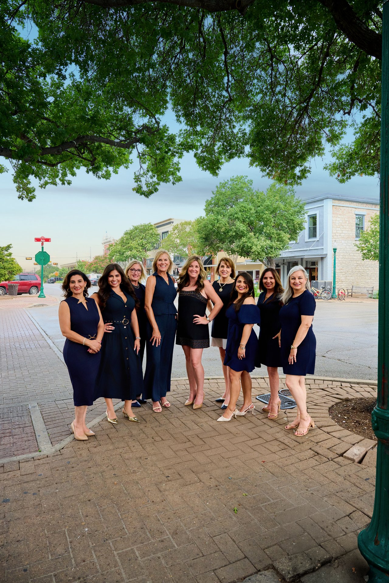 Nine women from a real estate team pose together on a brick sidewalk beneath leafy trees.