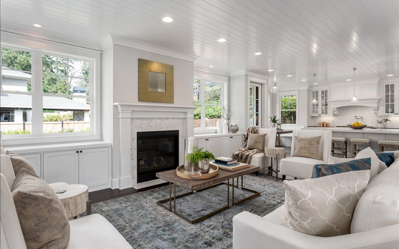 Bright living room with a white fireplace, large windows, and an adjoining kitchen.