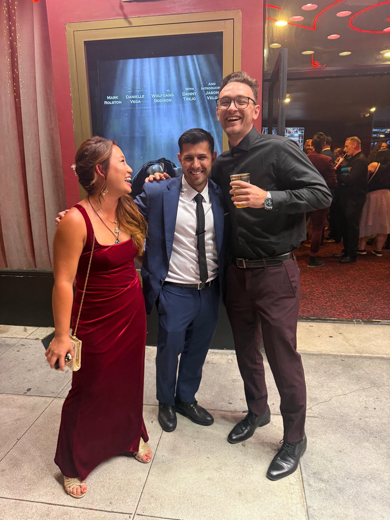 Three adults smiling together at an event, with the man on the right holding a drink.