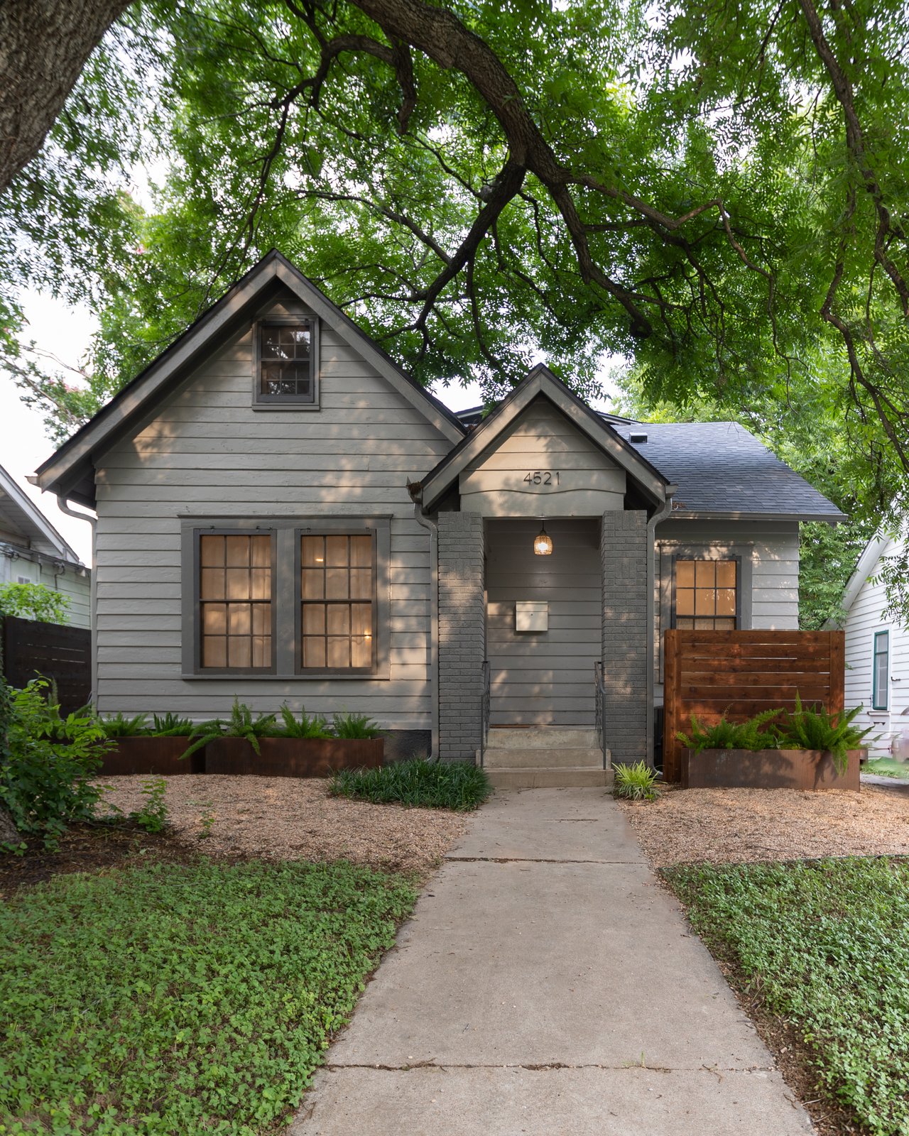 Gray gabled house with brick porch columns and a walkway beneath mature trees.