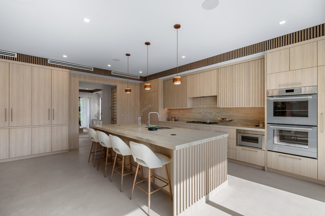 Modern kitchen with light wood cabinetry, a large island, bar stools, and double ovens.