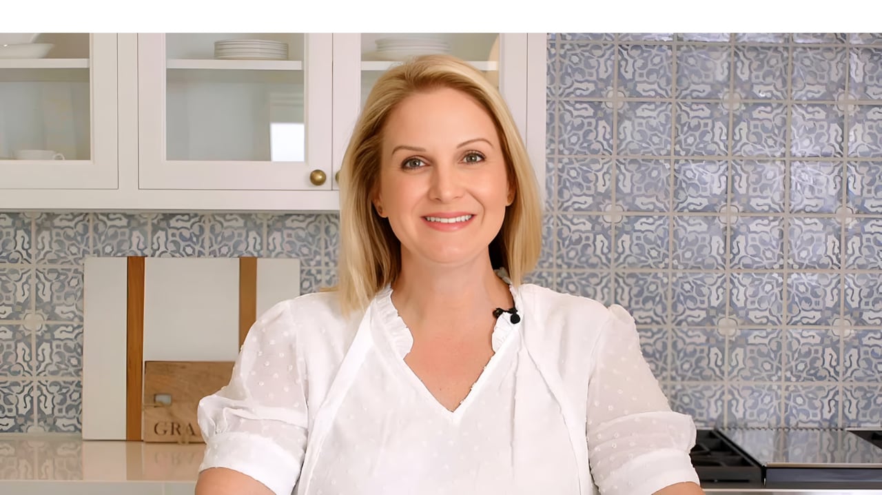 A professional headshot of Annie Clougherty smiling in a white blouse in a kitchen with blue and white tiled backsplash.