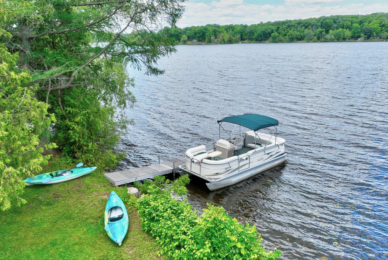 Wooded waterfront with a dock, pontoon boat, and two kayaks beside the lake.