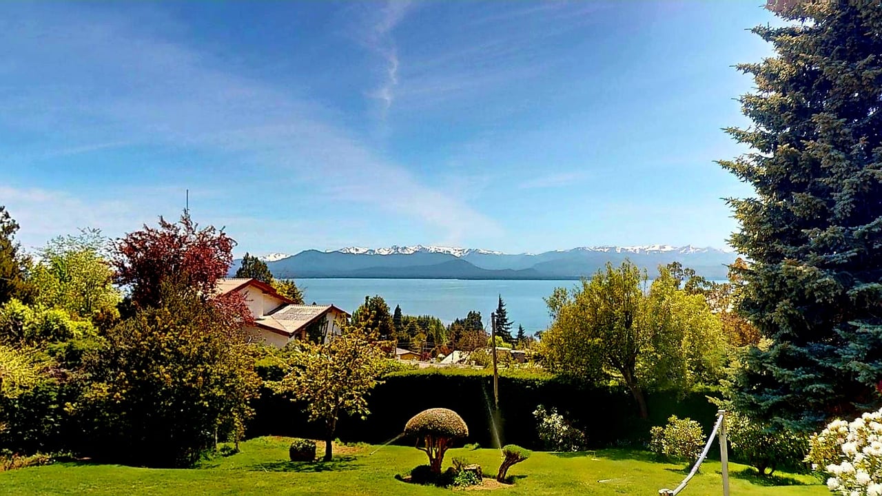 Landscaped garden overlooking a lake and snow-capped mountains, with part of a house among the trees.