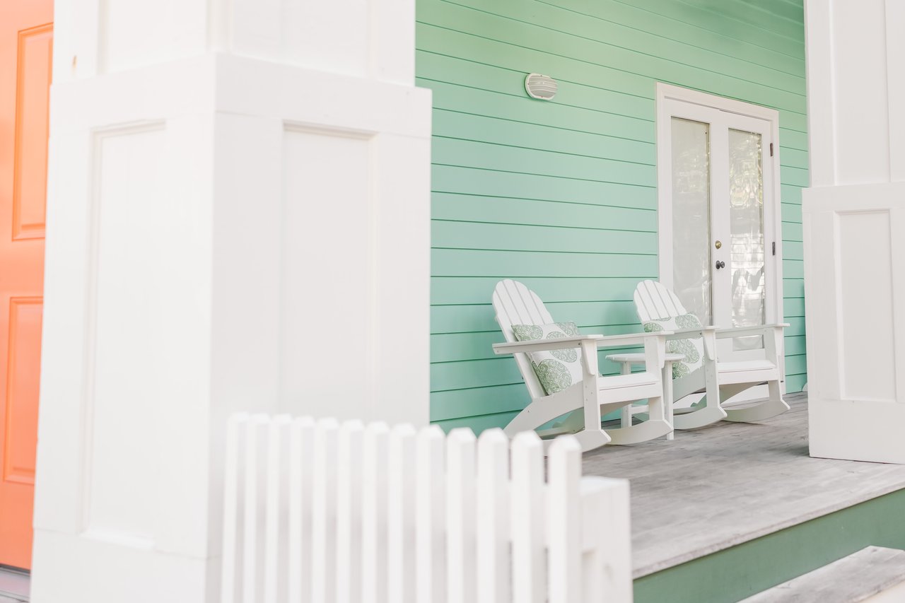 Sun-washed mint-green coastal cottage with a coral door, white rocking chairs and a picket fence at Watersound on 30A, Florida