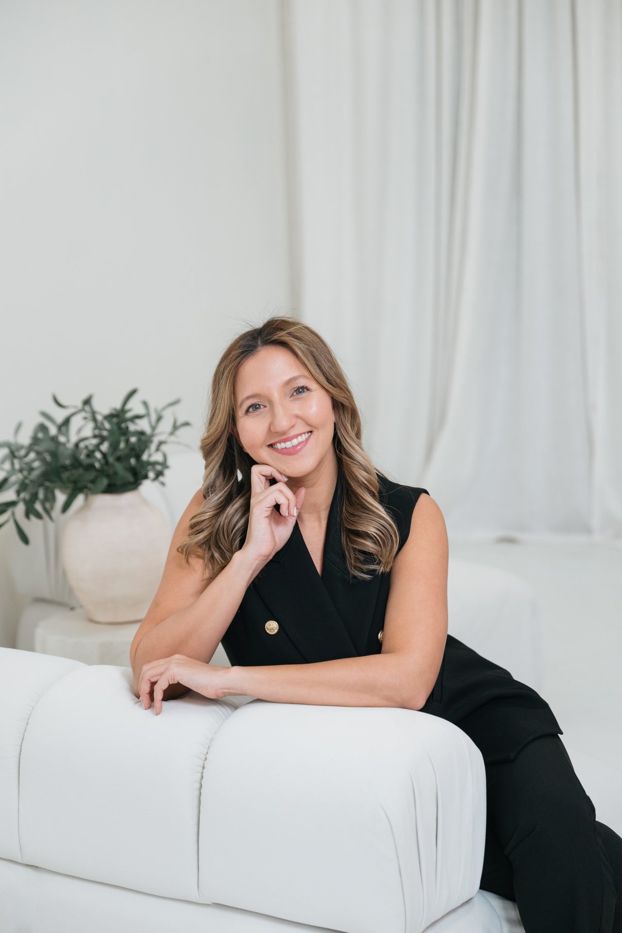 Simone De Los Angeles seated on a white sofa in a black sleeveless suit.