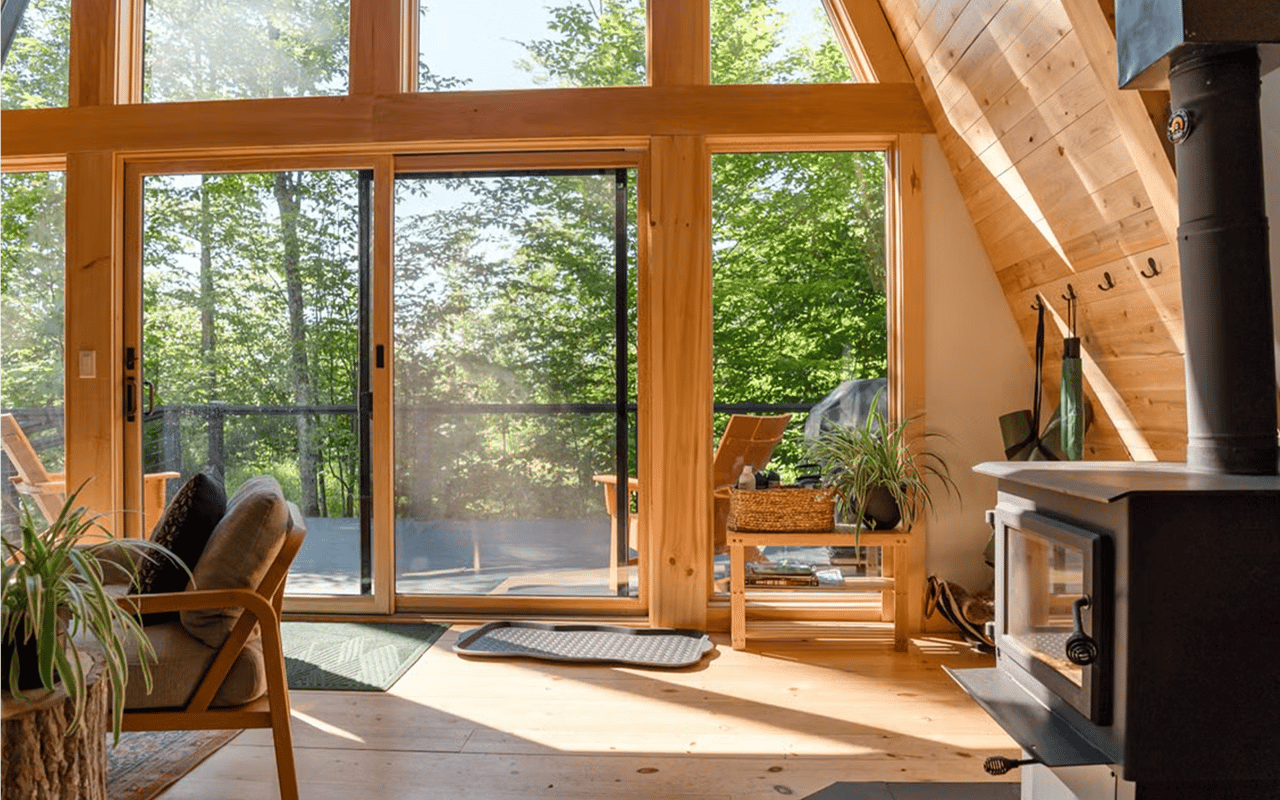 Cabin living room with a wood-burning stove and large windows overlooking trees.