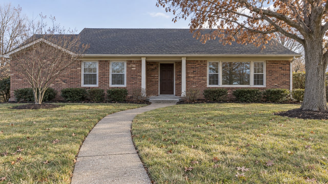 A modest red-brown brick home behind a deep lawn with a curving concrete walk and two deciduous trees in early winter.
