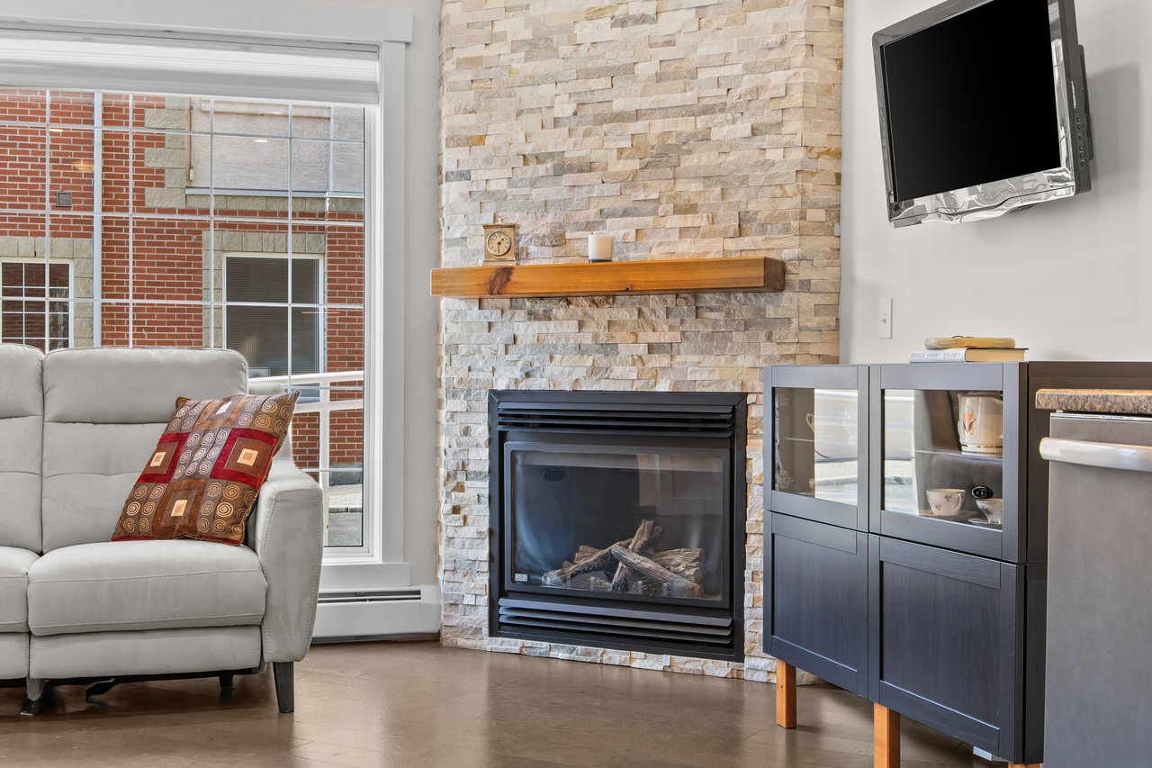 Living room with a stone fireplace, wooden mantel, gray sofa, and large window.