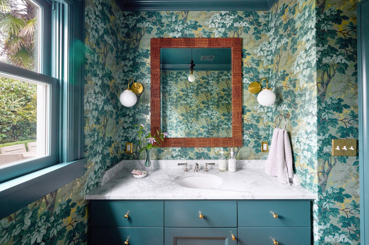 Bathroom vanity with teal cabinets, botanical wallpaper, marble countertop, and large framed mirror.
